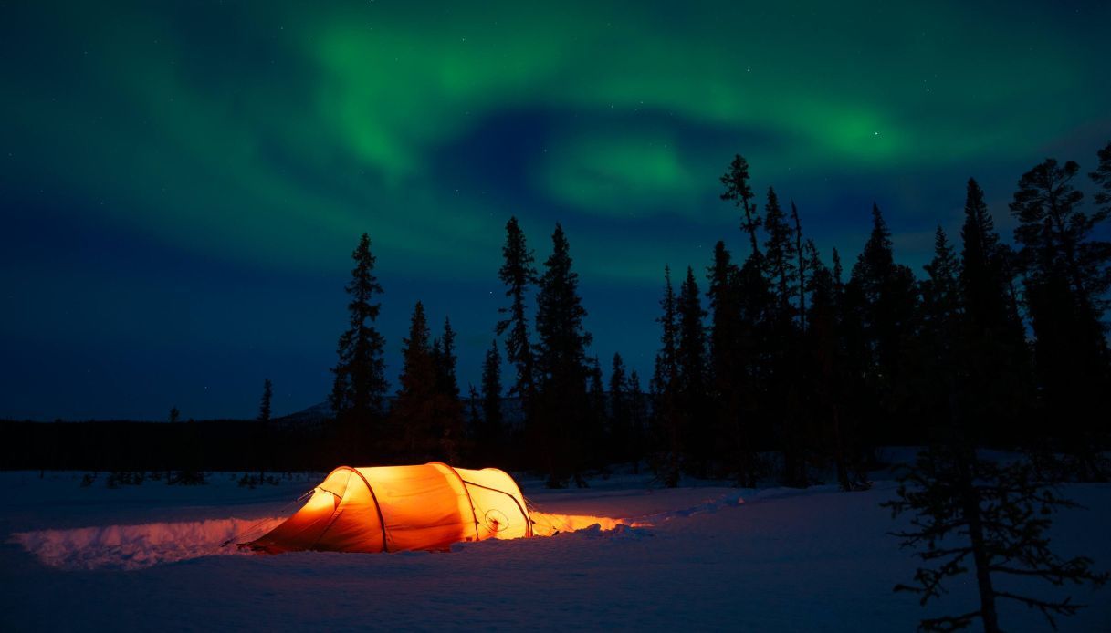 Una tenda illuminata dall'interno campeggia nella neve sotto un cielo con aurora boreale